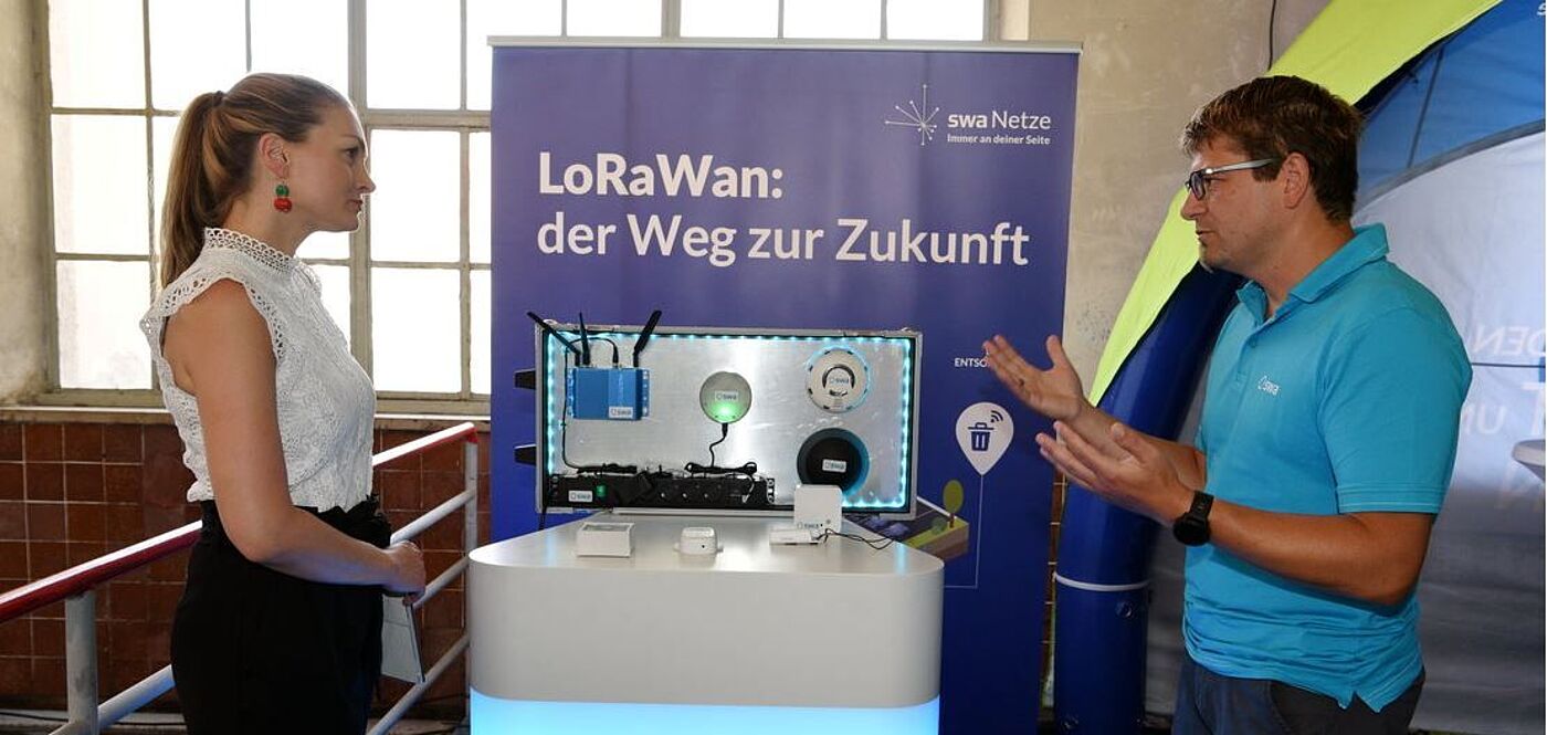 Zwei Personen stehen an einem Messestand mit einem beleuchteten Präsentationstisch, auf dem verschiedene technische Geräte und Sensoren ausgestellt sind. Im Hintergrund befindet sich ein blaues Banner mit der Aufschrift: ‚LoRaWan: der Weg zur Zukunft‘ sowie dem Logo der swa Netze. Die Szene zeigt eine Demonstration von LoRaWan-Technologie für intelligente Anwendungen.