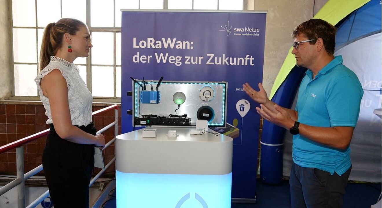 Zwei Personen stehen an einem Messestand mit einem beleuchteten Präsentationstisch, auf dem verschiedene technische Geräte und Sensoren ausgestellt sind. Im Hintergrund befindet sich ein blaues Banner mit der Aufschrift: ‚LoRaWan: der Weg zur Zukunft‘ sowie dem Logo der swa Netze. Die Szene zeigt eine Demonstration von LoRaWan-Technologie für intelligente Anwendungen.