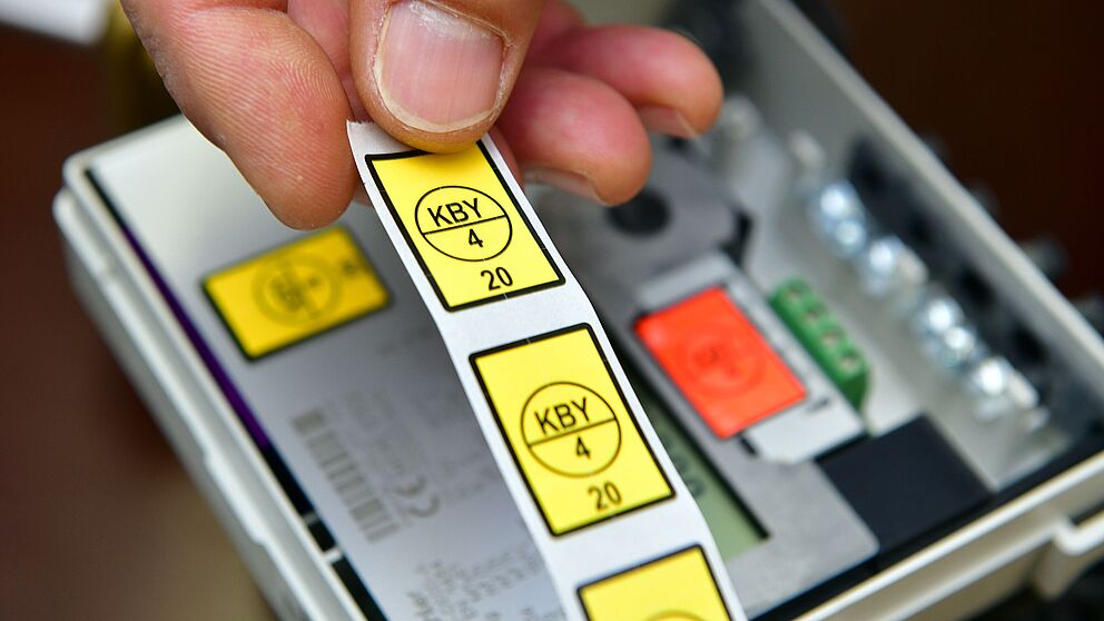 Nahaufnahme eines geöffneten Stromzählers. Im Vordergrund hält eine Hand einen Streifen mit gelben Prüfsiegeln, auf denen ‚KBY 4 20‘ steht. Im Hintergrund sind Teile des Zählers sichtbar, darunter eine orangefarbene Sicherung und grüne Anschlussklemmen.
