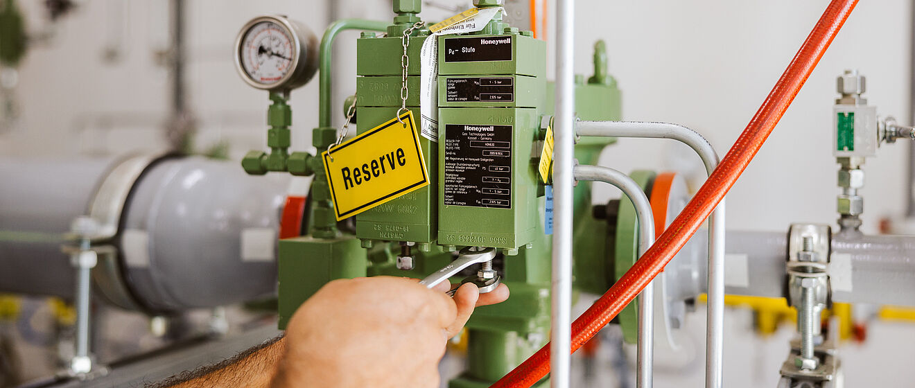 Nahaufnahme einer technischen Armatur mit grünem Steuergerät, zwei Manometern und einem gelben Schild mit der Aufschrift ‚Reserve‘. Sichtbar sind graue Rohrleitungen, rote Schläuche und metallische Verbindungen in einem hellen Technikraum.