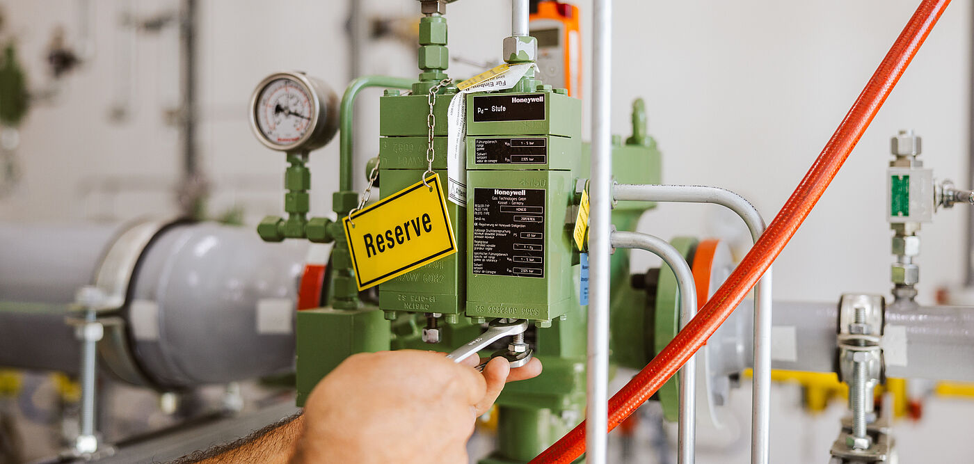 Nahaufnahme einer technischen Armatur mit grünem Steuergerät, zwei Manometern und einem gelben Schild mit der Aufschrift ‚Reserve‘. Sichtbar sind graue Rohrleitungen, rote Schläuche und metallische Verbindungen in einem hellen Technikraum.