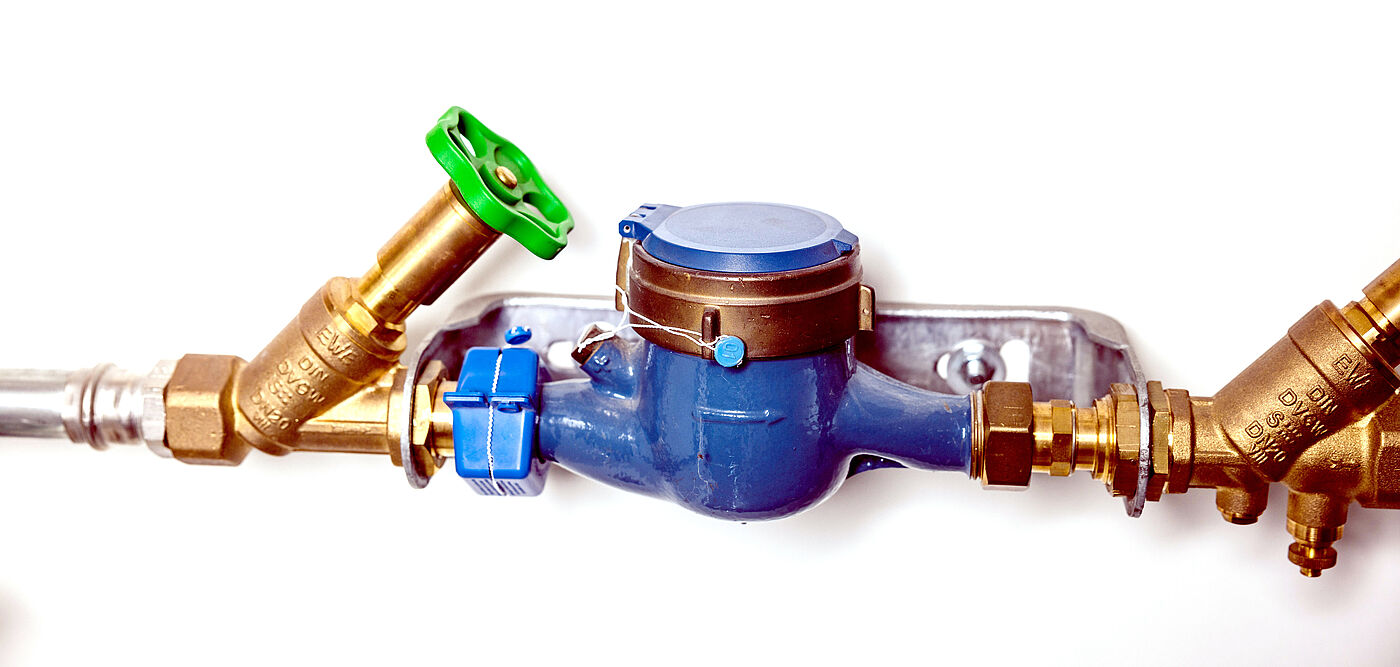 Ein Wasserzähler mit blauem Gehäuse ist in eine Rohrleitung eingebaut. Links befindet sich ein Messingventil mit grünem Handrad, daneben ein blaues Absperrventil. Rechts sind weitere Messingarmaturen und Anschlüsse sichtbar. Oberhalb des Zählers steht der Schriftzug ‚Wasserzähler‘.