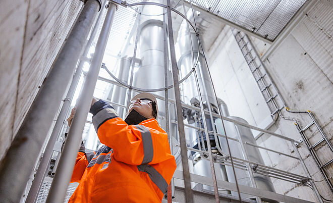 Arbeiter in auffälliger orangefarbener Warnschutzkleidung führt Wartungsarbeiten an einer metallischen Rohr- und Leitungskonstruktion in einem industriellen Schacht durch. Im Hintergrund sind große senkrechte Rohre, Gitterstrukturen und eine Leiter sichtbar, die auf eine technische Anlage oder Versorgungsinfrastruktur hinweisen. Verlinkung zur Wärme Seite.