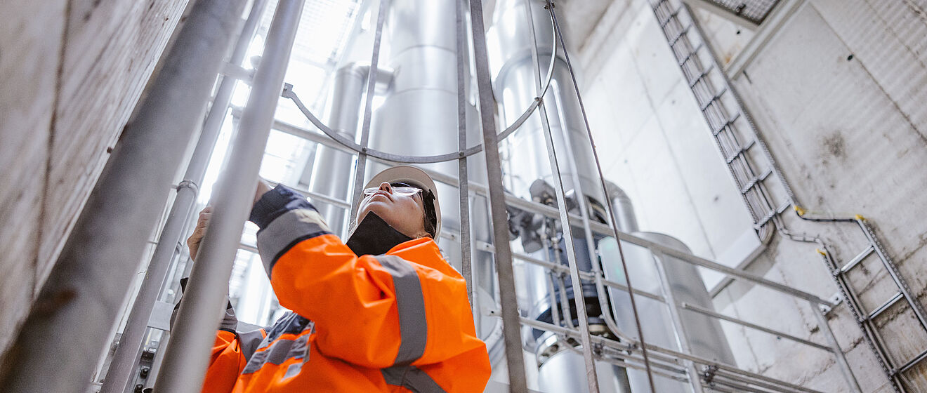 Arbeiter in auffälliger orangefarbener Warnschutzkleidung führt Wartungsarbeiten an einer metallischen Rohr- und Leitungskonstruktion in einem industriellen Schacht durch. Im Hintergrund sind große senkrechte Rohre, Gitterstrukturen und eine Leiter sichtbar, die auf eine technische Anlage oder Versorgungsinfrastruktur hinweisen. Verlinkung zur Wärme Seite.