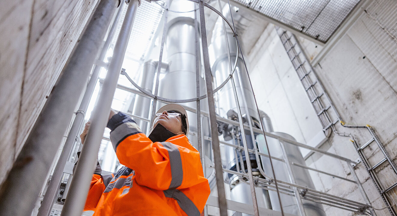 Arbeiter in auffälliger orangefarbener Warnschutzkleidung führt Wartungsarbeiten an einer metallischen Rohr- und Leitungskonstruktion in einem industriellen Schacht durch. Im Hintergrund sind große senkrechte Rohre, Gitterstrukturen und eine Leiter sichtbar, die auf eine technische Anlage oder Versorgungsinfrastruktur hinweisen. Verlinkung zur Wärme Seite.