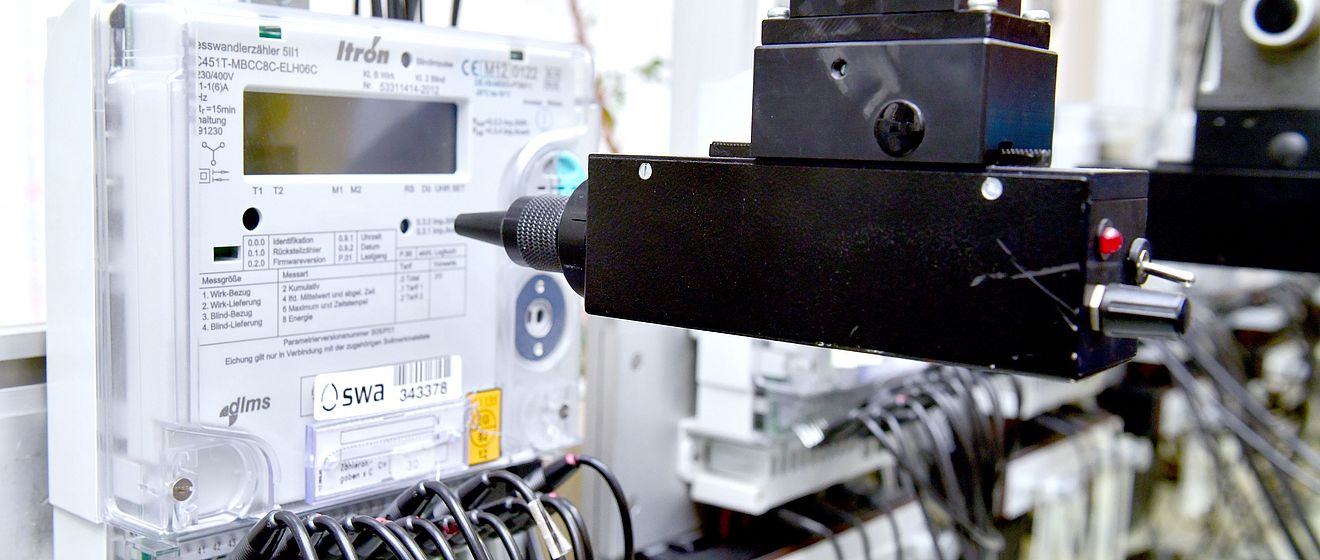 Nahaufnahme eines elektronischen Stromzählers mit dem Logo der swa Netze, montiert in einer Prüfvorrichtung. Der Zähler ist über zahlreiche schwarze Kabel verbunden, die nach unten führen. Vor dem Gerät befindet sich ein schwarzer Prüfkopf, der Teil einer automatisierten Testeinrichtung ist. Die Szene zeigt Messtechnik und Qualitätssicherung für intelligente Stromnetze.