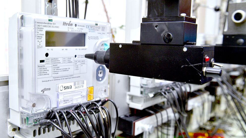 Nahaufnahme eines elektronischen Stromzählers mit dem Logo der swa Netze, montiert in einer Prüfvorrichtung. Der Zähler ist über zahlreiche schwarze Kabel verbunden, die nach unten führen. Vor dem Gerät befindet sich ein schwarzer Prüfkopf, der Teil einer automatisierten Testeinrichtung ist. Die Szene zeigt Messtechnik und Qualitätssicherung für intelligente Stromnetze.