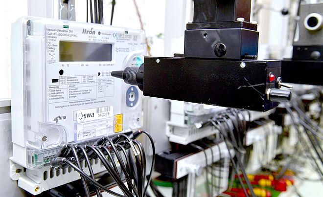 Nahaufnahme eines elektronischen Stromzählers mit dem Logo der swa Netze, montiert in einer Prüfvorrichtung. Der Zähler ist über zahlreiche schwarze Kabel verbunden, die nach unten führen. Vor dem Gerät befindet sich ein schwarzer Prüfkopf, der Teil einer automatisierten Testeinrichtung ist. Die Szene zeigt Messtechnik und Qualitätssicherung für intelligente Stromnetze.