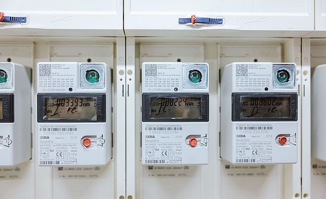 Nahaufnahme von drei digitalen Stromzählern, die in einer weißen Schalttafel installiert sind. Jeder Zähler zeigt ein Display mit Verbrauchswerten in Kilowattstunden, einen roten Bedientaster und einen QR-Code in der oberen Ecke. Die Geräte sind sauber angeordnet und beschriftet, was eine moderne Infrastruktur für Energie- und Verbrauchsmessung darstellt. Die Szene verdeutlicht den Einsatz von Smart-Meter-Technologie für präzise Stromabrechnung und Netzüberwachung.