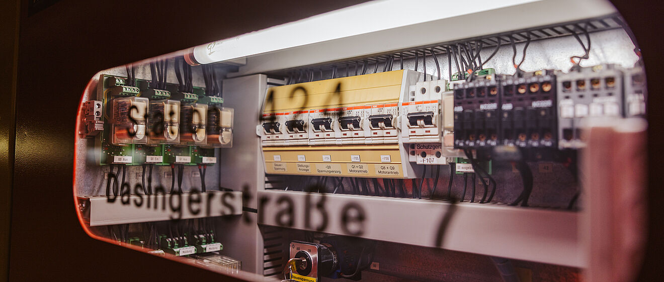 Detailansicht einer geöffneten Schaltanlage mit elektrischen Komponenten: Sichtbar sind mehrere Sicherungen, Schalter und Relais, die ordentlich in Reihen angeordnet sind. Schwarze Kabel verlaufen zu den Anschlüssen, darüber eine helle Beleuchtung. Auf der Scheibe ist die Beschriftung ‚Dasingerstraße‘ und eine Kennzeichnung ‚S421‘ erkennbar. Die Szene zeigt eine Steuer- und Verteilertechnik für Energieversorgung in einem Technikraum. Verlinkung zur Lastprofile Strom Seite.