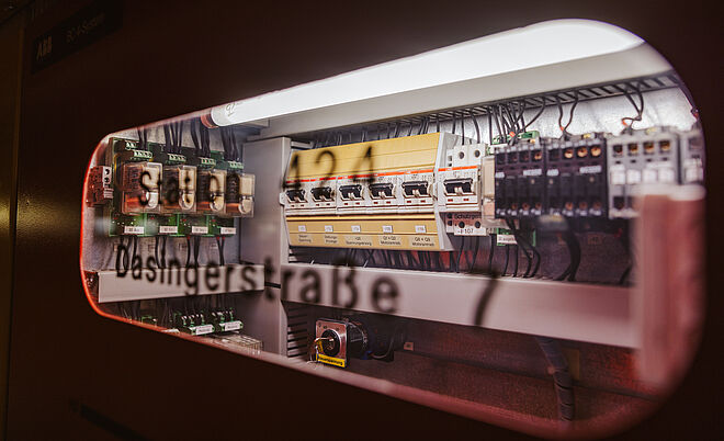 Detailansicht einer geöffneten Schaltanlage mit elektrischen Komponenten: Sichtbar sind mehrere Sicherungen, Schalter und Relais, die ordentlich in Reihen angeordnet sind. Schwarze Kabel verlaufen zu den Anschlüssen, darüber eine helle Beleuchtung. Auf der Scheibe ist die Beschriftung ‚Dasingerstraße‘ und eine Kennzeichnung ‚S421‘ erkennbar. Die Szene zeigt eine Steuer- und Verteilertechnik für Energieversorgung in einem Technikraum. Verlinkung zur Lastprofile Strom Seite.