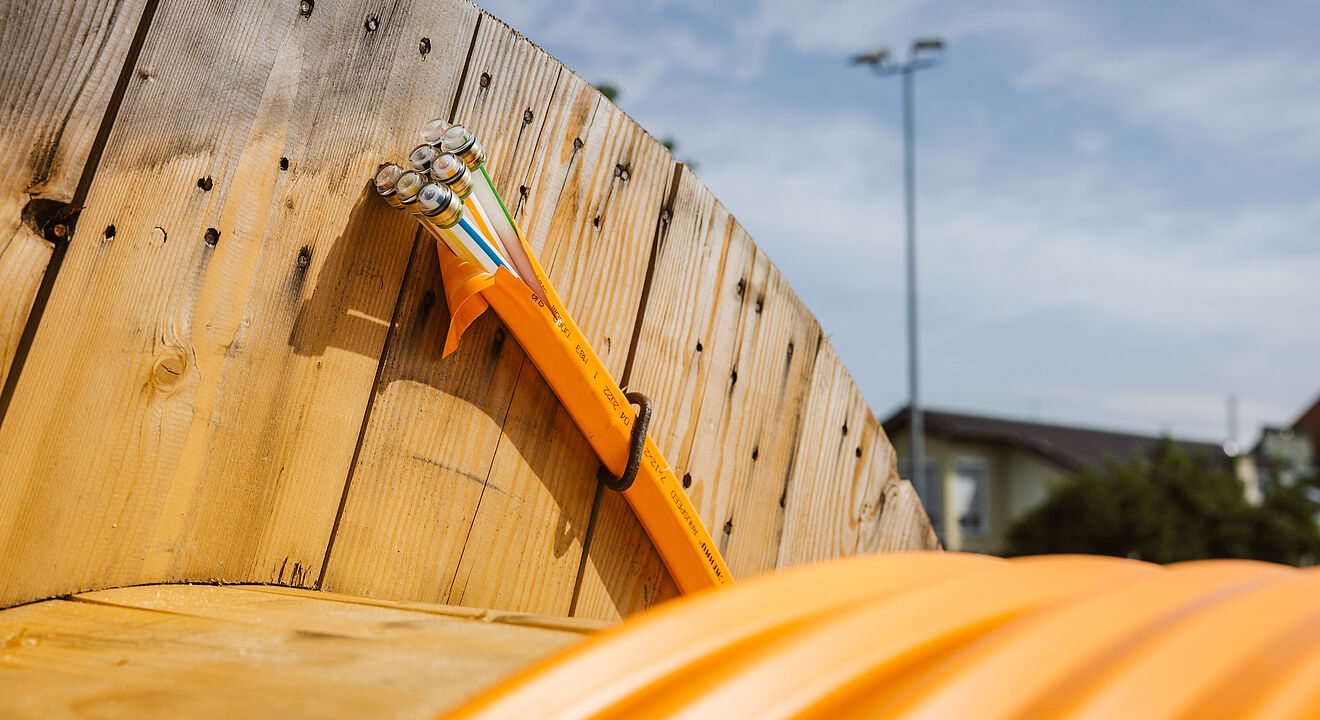 Nahaufnahme einer großen Holz-Kabeltrommel mit aufgewickeltem orangefarbenem Schutzrohr. Aus dem Rohr ragen mehrere Glasfaserkabel mit farbigen Markierungen heraus. Die Szene befindet sich im Freien bei klarem Himmel, im Hintergrund sind unscharf ein Gebäude und ein Lichtmast zu erkennen. Das Bild symbolisiert den Ausbau moderner Glasfaser- und Breitbandinfrastruktur. Verlinkung zur Kommunikation Seite.