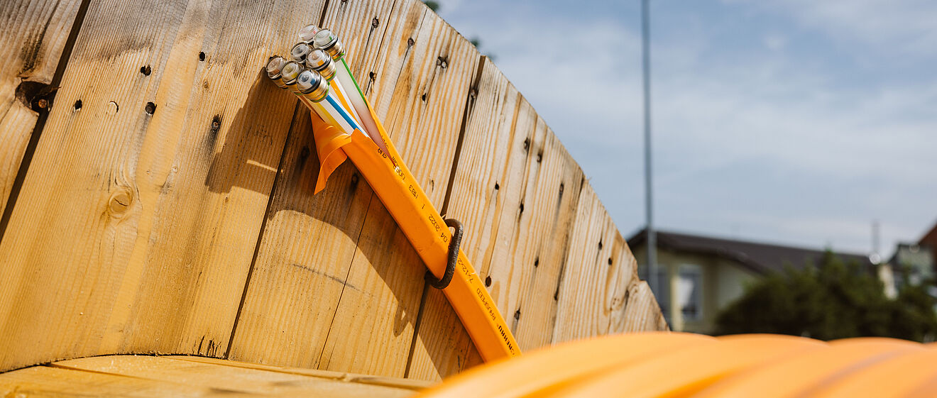 Nahaufnahme einer großen Holz-Kabeltrommel mit aufgewickeltem orangefarbenem Schutzrohr. Aus dem Rohr ragen mehrere Glasfaserkabel mit farbigen Markierungen heraus. Die Szene befindet sich im Freien bei klarem Himmel, im Hintergrund sind unscharf ein Gebäude und ein Lichtmast zu erkennen. Das Bild symbolisiert den Ausbau moderner Glasfaser- und Breitbandinfrastruktur. Verlinkung zur Kommunikation Seite.