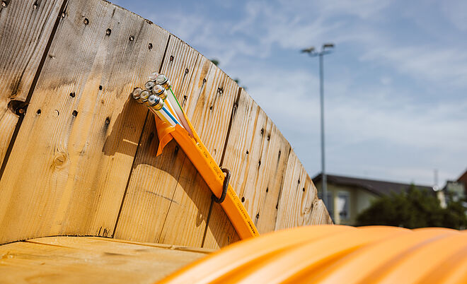 Nahaufnahme einer großen Holz-Kabeltrommel mit aufgewickeltem orangefarbenem Schutzrohr. Aus dem Rohr ragen mehrere Glasfaserkabel mit farbigen Markierungen heraus. Die Szene befindet sich im Freien bei klarem Himmel, im Hintergrund sind unscharf ein Gebäude und ein Lichtmast zu erkennen. Das Bild symbolisiert den Ausbau moderner Glasfaser- und Breitbandinfrastruktur. Verlinkung zur Kommunikation Seite.