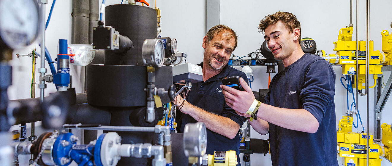 Zwei Personen arbeiten gemeinsam an einer komplexen Rohrleitungs- und Armaturenanlage in einem Technikraum. Sichtbar sind schwarze isolierte Rohre, mehrere Druckmessgeräte, Ventile und gelbe Sicherheitsmarkierungen. Im Hintergrund befinden sich weitere technische Komponenten, Kabel und Steuerungselemente. Verlinkung zur Einspeisung Gas Seite.