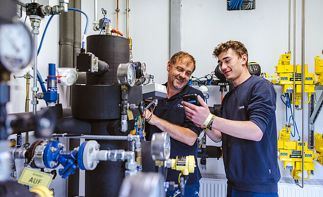 Zwei Personen arbeiten gemeinsam an einer komplexen Rohrleitungs- und Armaturenanlage in einem Technikraum. Sichtbar sind schwarze isolierte Rohre, mehrere Druckmessgeräte, Ventile und gelbe Sicherheitsmarkierungen. Im Hintergrund befinden sich weitere technische Komponenten, Kabel und Steuerungselemente. Verlinkung zur Einspeisung Gas Seite.