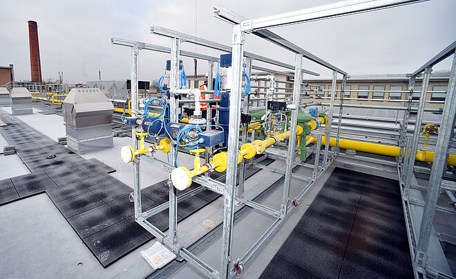 Auf einem Flachdach ist eine technische Anlage installiert, die aus mehreren Metallrahmen mit gelben Rohrleitungen und blauen Armaturen besteht. Die Konstruktion enthält Ventile, Messgeräte und Leitungen, die miteinander verbunden sind. Im Hintergrund sind weitere Gebäudeteile, Lüftungsanlagen und ein hoher Schornstein zu sehen. Der Himmel ist leicht bewölkt. Die Szene zeigt eine moderne Gas- oder Energieversorgungsinstallation auf einem Industrie- oder Betriebsgebäude. Verlinkung zur industriellen Anwendung Seite.