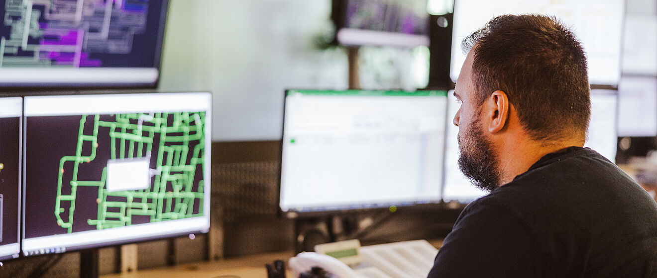Mitarbeiter überwacht Netzleitstelle an mehreren Monitoren: Auf den Bildschirmen sind komplexe Netzpläne und Steuerungssoftware mit grünen und violetten Leitungsdarstellungen sowie Datenfenstern sichtbar. Im Vordergrund befindet sich ein Schreibtisch mit Tastatur und Telefon. Die Szene zeigt die Überwachung und Steuerung von Energie- und Versorgungsnetzen in einem modernen Leitstand. Verlinkung zur Netzanschluss Strom Seite.