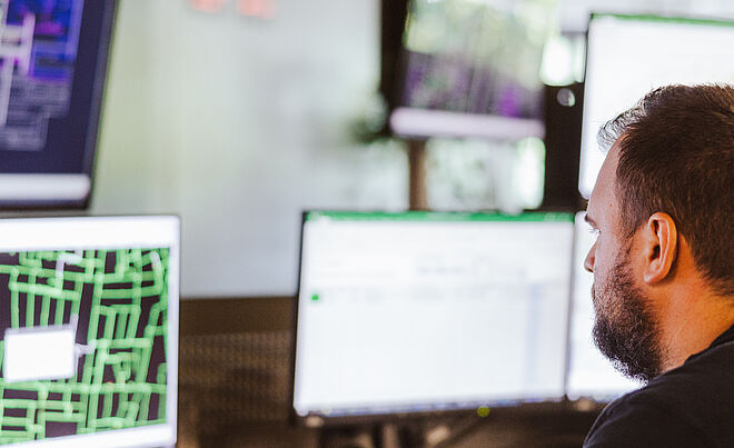 Mitarbeiter überwacht Netzleitstelle an mehreren Monitoren: Auf den Bildschirmen sind komplexe Netzpläne und Steuerungssoftware mit grünen und violetten Leitungsdarstellungen sowie Datenfenstern sichtbar. Im Vordergrund befindet sich ein Schreibtisch mit Tastatur und Telefon. Die Szene zeigt die Überwachung und Steuerung von Energie- und Versorgungsnetzen in einem modernen Leitstand. Verlinkung zur Netzanschluss Strom Seite.