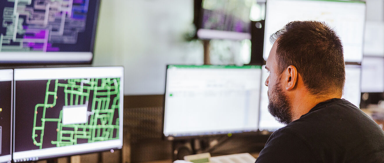 Mitarbeiter überwacht Netzleitstelle an mehreren Monitoren: Auf den Bildschirmen sind komplexe Netzpläne und Steuerungssoftware mit grünen und violetten Leitungsdarstellungen sowie Datenfenstern sichtbar. Im Vordergrund befindet sich ein Schreibtisch mit Tastatur und Telefon. Die Szene zeigt die Überwachung und Steuerung von Energie- und Versorgungsnetzen in einem modernen Leitstand. Verlinkung zur Netzanschluss Strom Seite.