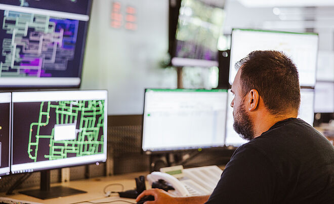 Mitarbeiter überwacht Netzleitstelle an mehreren Monitoren: Auf den Bildschirmen sind komplexe Netzpläne und Steuerungssoftware mit grünen und violetten Leitungsdarstellungen sowie Datenfenstern sichtbar. Im Vordergrund befindet sich ein Schreibtisch mit Tastatur und Telefon. Die Szene zeigt die Überwachung und Steuerung von Energie- und Versorgungsnetzen in einem modernen Leitstand. Verlinkung zur Netzanschluss Strom Seite.