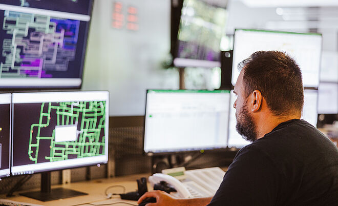 Mitarbeiter überwacht Netzleitstelle an mehreren Monitoren: Auf den Bildschirmen sind komplexe Netzpläne und Steuerungssoftware mit grünen und violetten Leitungsdarstellungen sowie Datenfenstern sichtbar. Im Vordergrund befindet sich ein Schreibtisch mit Tastatur und Telefon. Die Szene zeigt die Überwachung und Steuerung von Energie- und Versorgungsnetzen in einem modernen Leitstand. Verlinkung zur Netzanschluss Strom Seite.