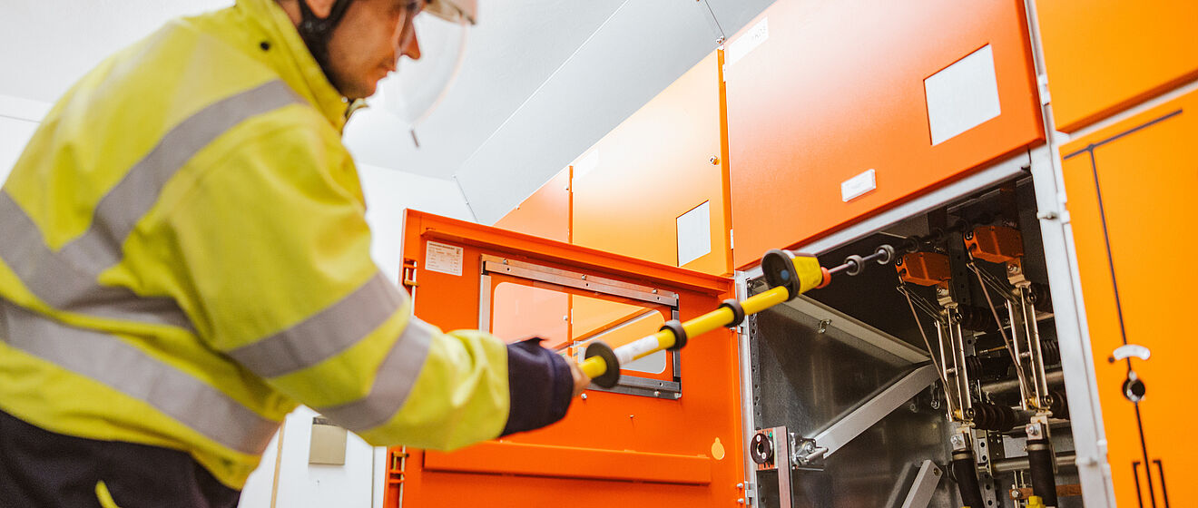 Elektrofachkraft arbeitet an einer Schaltanlage: Die Person trägt eine gelbe Warnschutzjacke und einen weißen Schutzhelm und bedient mit einer isolierten Schaltstange eine geöffnete orangefarbene Mittelspannungs-Schaltanlage. Im Hintergrund sind weitere geschlossene Schaltschränke zu sehen. Die Szene zeigt sicheres Arbeiten an elektrischen Anlagen im Innenbereich. Verlinkung zur Strom Seite.