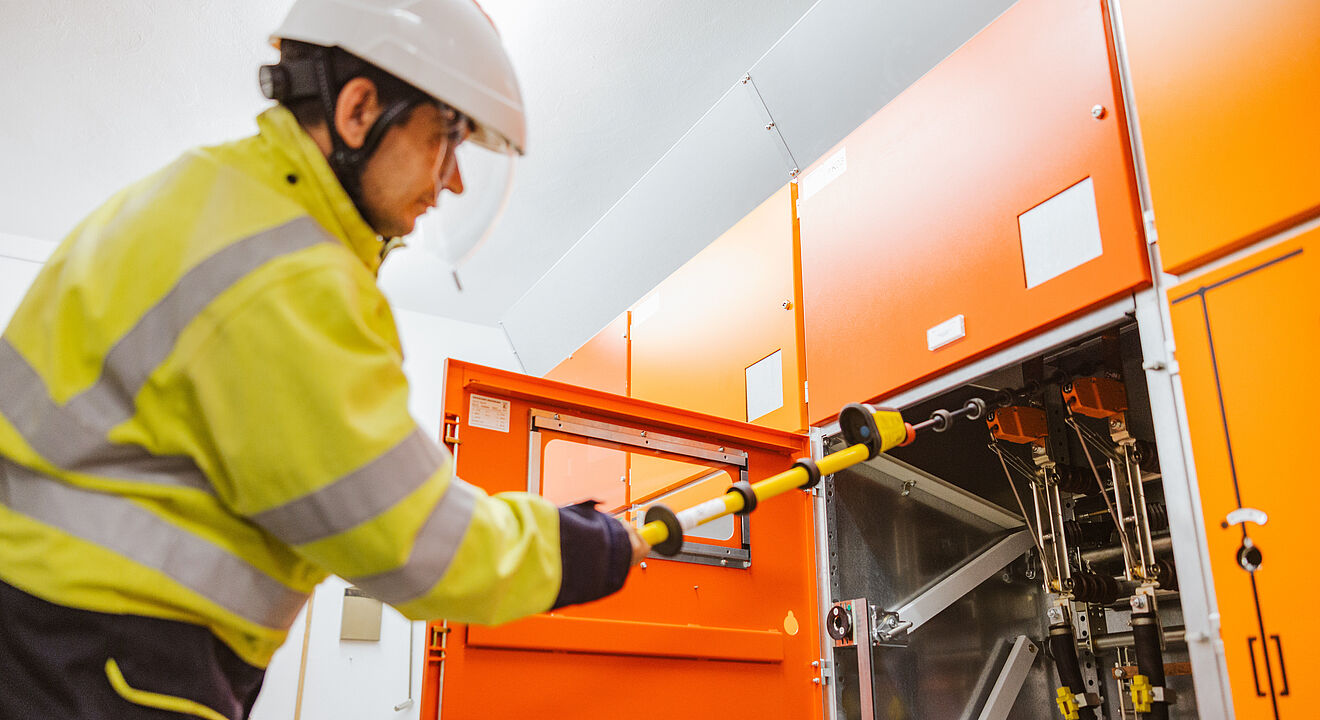 Elektrofachkraft arbeitet an einer Schaltanlage: Die Person trägt eine gelbe Warnschutzjacke und einen weißen Schutzhelm und bedient mit einer isolierten Schaltstange eine geöffnete orangefarbene Mittelspannungs-Schaltanlage. Im Hintergrund sind weitere geschlossene Schaltschränke zu sehen. Die Szene zeigt sicheres Arbeiten an elektrischen Anlagen im Innenbereich. Verlinkung zur Strom Seite.