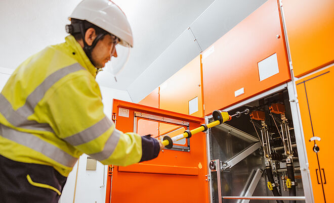 Elektrofachkraft arbeitet an einer Schaltanlage: Die Person trägt eine gelbe Warnschutzjacke und einen weißen Schutzhelm und bedient mit einer isolierten Schaltstange eine geöffnete orangefarbene Mittelspannungs-Schaltanlage. Im Hintergrund sind weitere geschlossene Schaltschränke zu sehen. Die Szene zeigt sicheres Arbeiten an elektrischen Anlagen im Innenbereich. Verlinkung zur Strom Seite.
