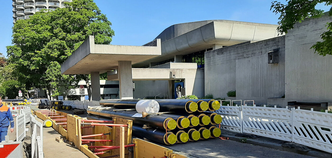 Baustelle für Rohrleitungsarbeiten im städtischen Bereich: Im Vordergrund sind gelbe Baugrubenverbauungen mit roten Streben zu sehen, dahinter liegen mehrere Bündel schwarzer Rohre mit gelben Endkappen. Die Umgebung zeigt moderne Betonarchitektur, weiße Absperrgitter und einen hohen Gebäudeturm im Hintergrund. Bäume spenden Schatten, die Szene ist bei sonnigem Wetter aufgenommen.