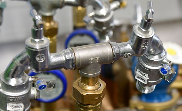 Mehrere Wasserzähler und Armaturen in einer Installationseinheit. Im Vordergrund ist ein Wasserzähler mit blauem Ring und digitalem Display sichtbar, montiert an einem Messingrohr mit Ventilen. Weitere Zähler und Rohrverbindungen befinden sich im Hintergrund. Die Armaturen sind aus Metall, teilweise mit Prüfplaketten wie ‚SWA 2020‘ versehen. Verlinkung zur Standrohr-Wasserzähler Seite.