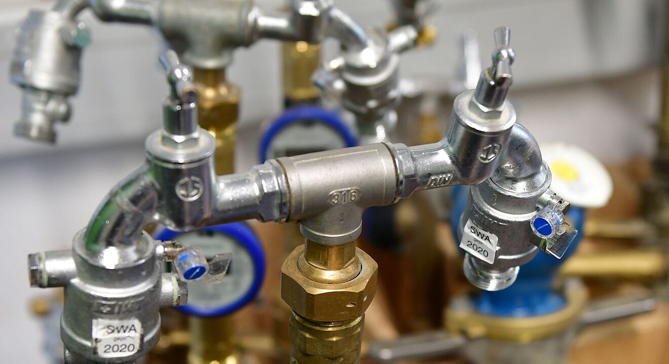 Mehrere Wasserzähler und Armaturen in einer Installationseinheit. Im Vordergrund ist ein Wasserzähler mit blauem Ring und digitalem Display sichtbar, montiert an einem Messingrohr mit Ventilen. Weitere Zähler und Rohrverbindungen befinden sich im Hintergrund. Die Armaturen sind aus Metall, teilweise mit Prüfplaketten wie ‚SWA 2020‘ versehen. Verlinkung zur Standrohr-Wasserzähler Seite.