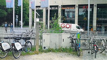Fahrräder stehen an einer öffentlich zugänglichen Ladeeinrichtung neben einem hellen Technikschrank. Die Anlage befindet sich auf einer geschotterten Fläche mit Gras und Wildpflanzen. Hinter der Installation verlaufen ein Metallzaun und eine Straße; im Hintergrund sind Bäume und ein Transportfahrzeug erkennbar.