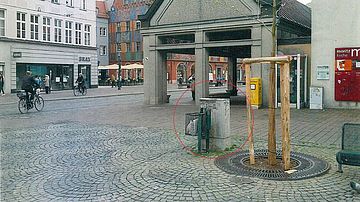Städtischer Platz mit Pflasterbelag vor einer offenen Markthalle und mehrgeschossigen Gebäuden. Im Vordergrund steht ein junger Baum mit Baumscheibe; daneben befindet sich ein grau gestrichenes Technikgehäuse mit angebrachtem Abfallkorb, das durch einen roten Kreis hervorgehoben ist. Im Hintergrund sind Sitzgelegenheiten, Fahrräder, Passanten sowie Geschäfts- und Kirchengebäude sichtbar. Die Markierung lenkt die Aufmerksamkeit auf den Standort eines technischen Objekts im öffentlichen Raum.