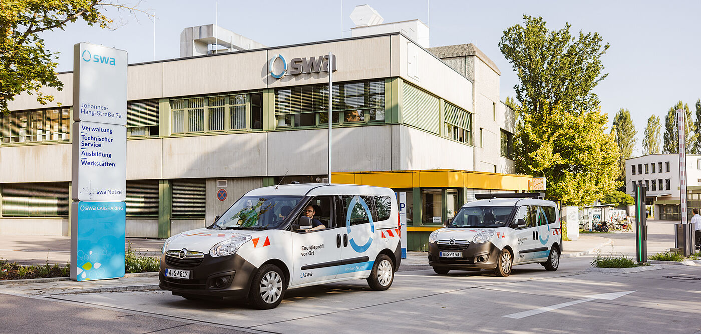 Vor einem modernen Verwaltungsgebäude mit der Aufschrift „SMI“ stehen zwei Fahrzeuge der Stadtwerke Augsburg (swa). Beide Fahrzeuge sind weiß mit blauen und roten grafischen Elementen und tragen die Beschriftung „Energie“ sowie das swa-Logo. Links im Bild befindet sich eine große Infotafel mit der Adresse „Johannes-Haag-Straße 7“ und Hinweisen zu Verwaltung, Technik, Service, Ausbildung und Werkstätten. Im Hintergrund sind Bäume und weitere Gebäude zu sehen, die Szene spielt bei Tageslicht. Verlinkung zur Unternehmen Seite.