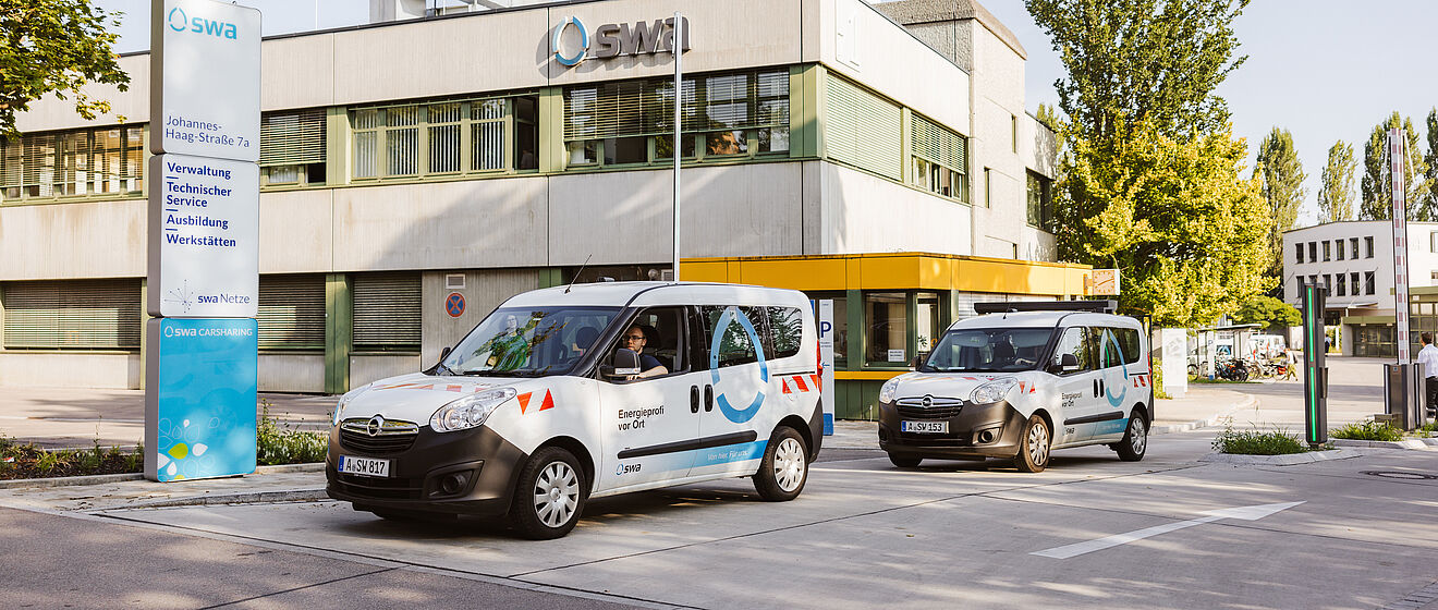 Vor einem modernen Verwaltungsgebäude mit der Aufschrift „SMI“ stehen zwei Fahrzeuge der Stadtwerke Augsburg (swa). Beide Fahrzeuge sind weiß mit blauen und roten grafischen Elementen und tragen die Beschriftung „Energie“ sowie das swa-Logo. Links im Bild befindet sich eine große Infotafel mit der Adresse „Johannes-Haag-Straße 7“ und Hinweisen zu Verwaltung, Technik, Service, Ausbildung und Werkstätten. Im Hintergrund sind Bäume und weitere Gebäude zu sehen, die Szene spielt bei Tageslicht. Verlinkung zur Unternehmen Seite.