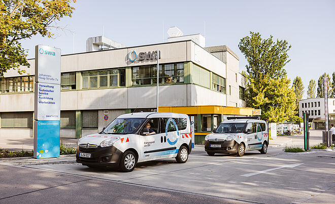Vor einem modernen Verwaltungsgebäude mit der Aufschrift „SMI“ stehen zwei Fahrzeuge der Stadtwerke Augsburg (swa). Beide Fahrzeuge sind weiß mit blauen und roten grafischen Elementen und tragen die Beschriftung „Energie“ sowie das swa-Logo. Links im Bild befindet sich eine große Infotafel mit der Adresse „Johannes-Haag-Straße 7“ und Hinweisen zu Verwaltung, Technik, Service, Ausbildung und Werkstätten. Im Hintergrund sind Bäume und weitere Gebäude zu sehen, die Szene spielt bei Tageslicht. Verlinkung zur Unternehmen Seite.