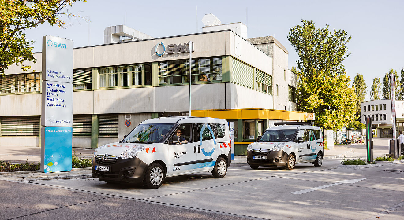 Vor einem modernen Verwaltungsgebäude mit der Aufschrift „SMI“ stehen zwei Fahrzeuge der Stadtwerke Augsburg (swa). Beide Fahrzeuge sind weiß mit blauen und roten grafischen Elementen und tragen die Beschriftung „Energie“ sowie das swa-Logo. Links im Bild befindet sich eine große Infotafel mit der Adresse „Johannes-Haag-Straße 7“ und Hinweisen zu Verwaltung, Technik, Service, Ausbildung und Werkstätten. Im Hintergrund sind Bäume und weitere Gebäude zu sehen, die Szene spielt bei Tageslicht. Verlinkung zur Unternehmen Seite.