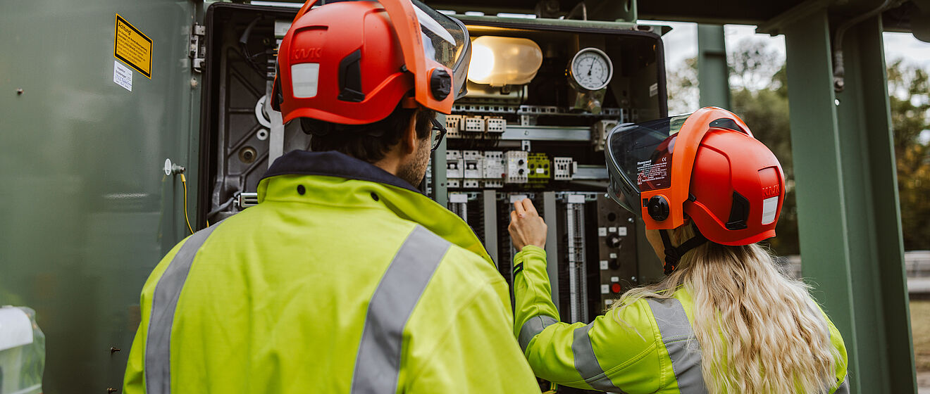 Zwei Personen in Warnschutzkleidung und roten Schutzhelmen arbeiten an einer geöffneten Schaltanlage in einem Umspannwerk. Eine Person hält ein Werkzeug und führt eine Einstellung an elektrischen Komponenten durch, während die andere daneben steht. Im Inneren der Anlage sind Schalter, Kabel, Messgeräte und ein Manometer sichtbar, darüber eine Lampe zur Beleuchtung. Die Szene zeigt Wartungsarbeiten an Hochspannungsanlagen im Bereich Stromversorgung und Netztechnik. Verlinkung zur §14a Energiewirtschaftsgesetz (EnWG) Seite.