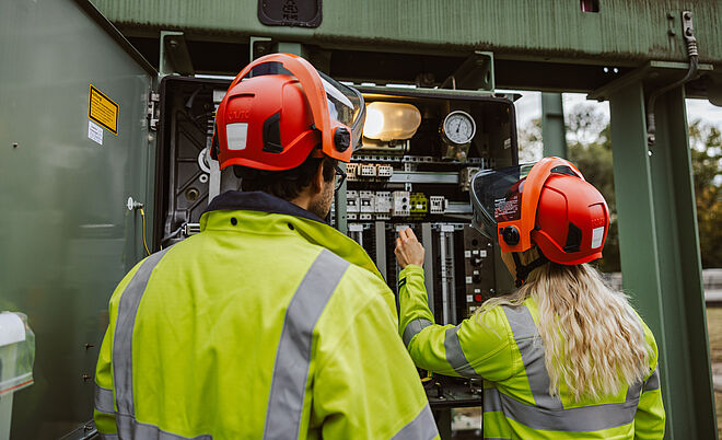 Zwei Personen in Warnschutzkleidung und roten Schutzhelmen arbeiten an einer geöffneten Schaltanlage in einem Umspannwerk. Eine Person hält ein Werkzeug und führt eine Einstellung an elektrischen Komponenten durch, während die andere daneben steht. Im Inneren der Anlage sind Schalter, Kabel, Messgeräte und ein Manometer sichtbar, darüber eine Lampe zur Beleuchtung. Die Szene zeigt Wartungsarbeiten an Hochspannungsanlagen im Bereich Stromversorgung und Netztechnik. Verlinkung zur §14a Energiewirtschaftsgesetz (EnWG) Seite.