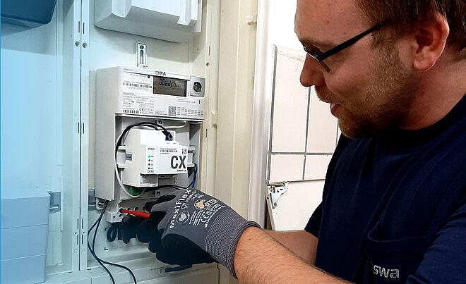 Ein Elektriker arbeitet an einem geöffneten Stromzählerkasten. Im Inneren ist ein moderner digitaler Stromzähler mit einem zusätzlichen Modul, das mit ‚CX‘ beschriftet ist, sichtbar. Der Elektriker trägt Arbeitshandschuhe und hält ein Werkzeug, um Kabel im unteren Bereich des Zählerkastens anzuschließen. Links und rechts sind weitere Komponenten des Schaltschrankes erkennbar. Verlinkung zur Messstellenbetrieb Strom Seite.