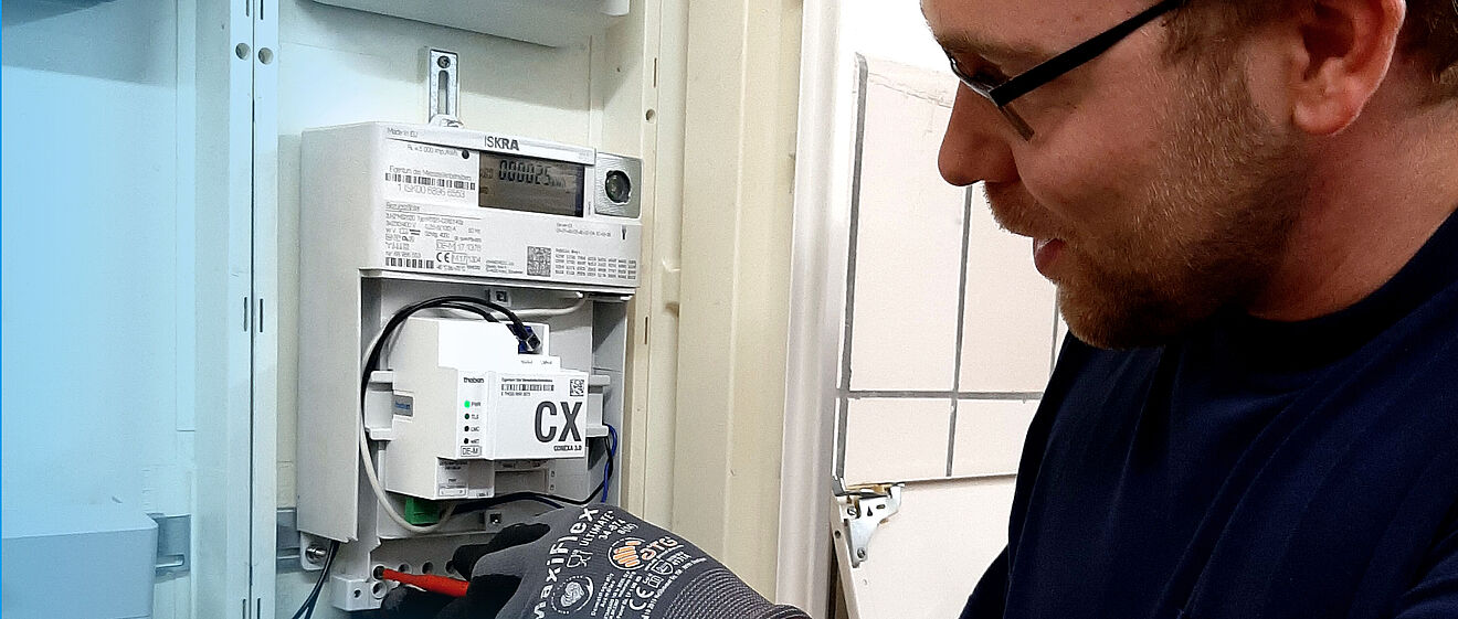 Ein Elektriker arbeitet an einem geöffneten Stromzählerkasten. Im Inneren ist ein moderner digitaler Stromzähler mit einem zusätzlichen Modul, das mit ‚CX‘ beschriftet ist, sichtbar. Der Elektriker trägt Arbeitshandschuhe und hält ein Werkzeug, um Kabel im unteren Bereich des Zählerkastens anzuschließen. Links und rechts sind weitere Komponenten des Schaltschrankes erkennbar. Verlinkung zur Messstellenbetrieb Strom Seite.