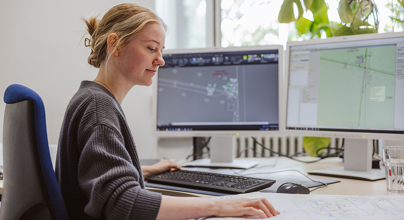 Eine Person sitzt an einem Schreibtisch und arbeitet an zwei großen Monitoren. Auf den Bildschirmen sind technische Pläne und CAD-Zeichnungen zu sehen. Die Person bedient die Tastatur und hat eine Hand auf einem großformatigen Papierplan, der auf dem Schreibtisch ausgebreitet liegt. Neben der Tastatur steht ein Glas Wasser. Im Hintergrund befindet sich ein Fenster mit Tageslicht und eine grüne Pflanze.