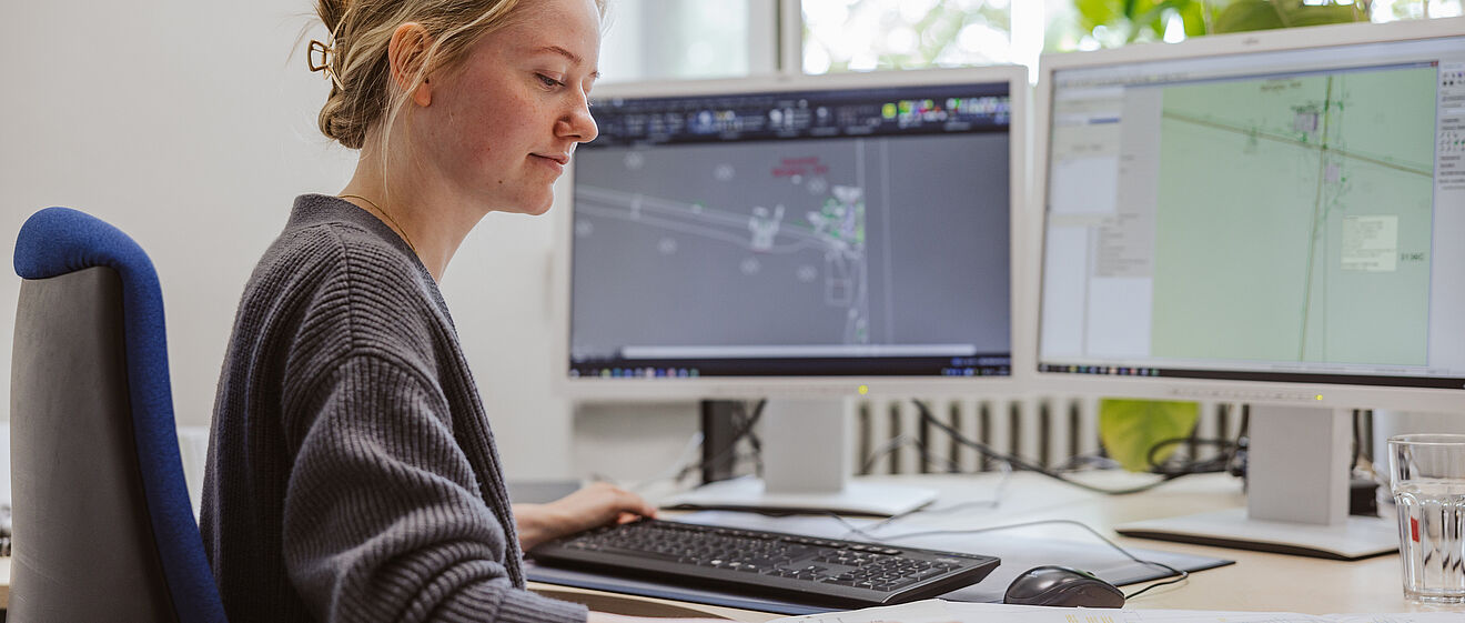 Eine Person sitzt an einem Schreibtisch und arbeitet an zwei großen Monitoren. Auf den Bildschirmen sind technische Pläne und CAD-Zeichnungen zu sehen. Die Person bedient die Tastatur und hat eine Hand auf einem großformatigen Papierplan, der auf dem Schreibtisch ausgebreitet liegt. Neben der Tastatur steht ein Glas Wasser. Im Hintergrund befindet sich ein Fenster mit Tageslicht und eine grüne Pflanze.