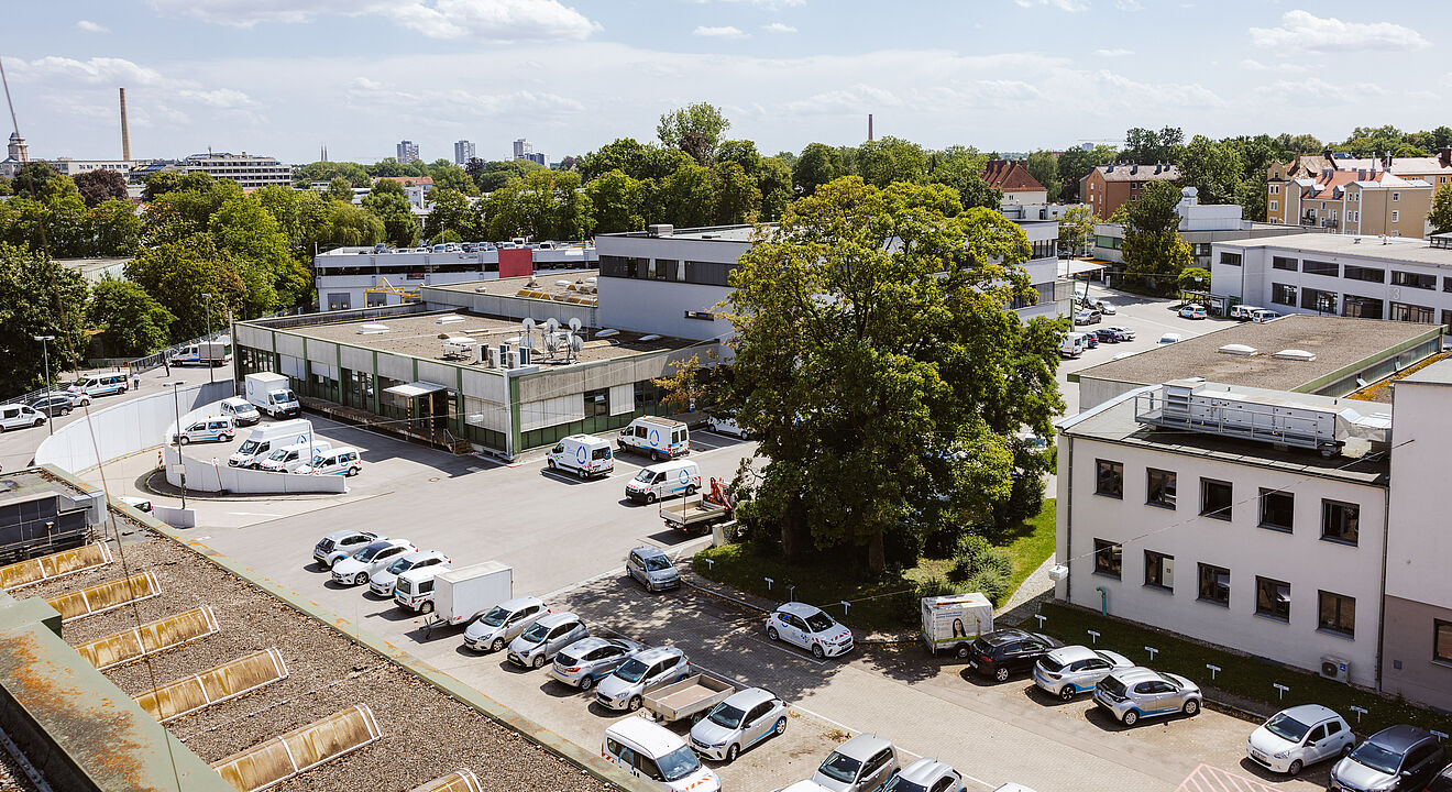 Luftaufnahme des Betriebsgeländes der swa Netze GmbH in Augsburg. Im Vordergrund sind mehrere geparkte Servicefahrzeuge und Pkw auf einem Parkplatz zu sehen. Dahinter befinden sich verschiedene Büro- und Werkstattgebäude, umgeben von Bäumen und Grünflächen. Im Hintergrund erstreckt sich die Stadt mit Wohnhäusern und Industriegebäuden, unter einem sonnigen Himmel mit vereinzelten Wolken. Die Szene vermittelt einen Überblick über die Infrastruktur und den Standort des Unternehmens.