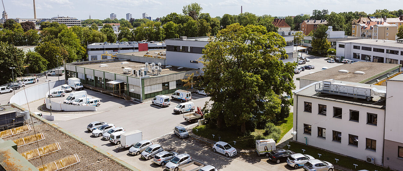 Luftaufnahme des Betriebsgeländes der swa Netze GmbH in Augsburg. Im Vordergrund sind mehrere geparkte Servicefahrzeuge und Pkw auf einem Parkplatz zu sehen. Dahinter befinden sich verschiedene Büro- und Werkstattgebäude, umgeben von Bäumen und Grünflächen. Im Hintergrund erstreckt sich die Stadt mit Wohnhäusern und Industriegebäuden, unter einem sonnigen Himmel mit vereinzelten Wolken. Die Szene vermittelt einen Überblick über die Infrastruktur und den Standort des Unternehmens.