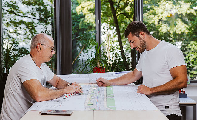 Zwei Personen sitzen an einem großen Tisch und betrachten technische Baupläne. Auf dem Tisch liegen mehrere großformatige Pläne mit detaillierten Zeichnungen und Markierungen. Im Hintergrund sind große Fenster mit Blick ins Grüne sowie eine rote Pflanze auf der Fensterbank sichtbar. Die Szene spielt in einem hellen Büro oder Besprechungsraum. Verlinkung zur Aufmassportal Seite.