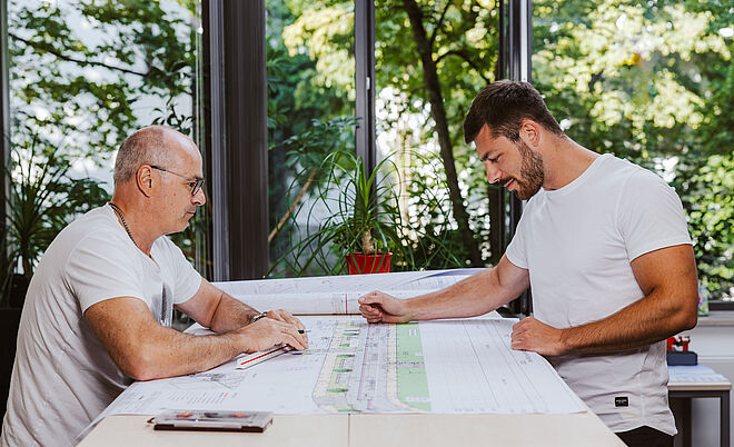 Zwei Personen sitzen an einem großen Tisch und betrachten technische Baupläne. Auf dem Tisch liegen mehrere großformatige Pläne mit detaillierten Zeichnungen und Markierungen. Im Hintergrund sind große Fenster mit Blick ins Grüne sowie eine rote Pflanze auf der Fensterbank sichtbar. Die Szene spielt in einem hellen Büro oder Besprechungsraum. Verlinkung zur Aufmassportal Seite.