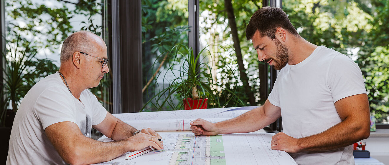 Zwei Personen sitzen an einem großen Tisch und betrachten technische Baupläne. Auf dem Tisch liegen mehrere großformatige Pläne mit detaillierten Zeichnungen und Markierungen. Im Hintergrund sind große Fenster mit Blick ins Grüne sowie eine rote Pflanze auf der Fensterbank sichtbar. Die Szene spielt in einem hellen Büro oder Besprechungsraum. Verlinkung zur Aufmassportal Seite.