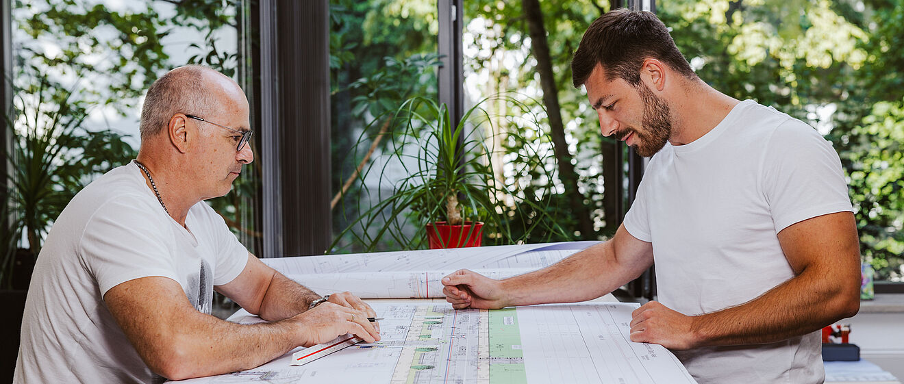 Zwei Personen sitzen an einem großen Tisch und betrachten technische Baupläne. Auf dem Tisch liegen mehrere großformatige Pläne mit detaillierten Zeichnungen und Markierungen. Im Hintergrund sind große Fenster mit Blick ins Grüne sowie eine rote Pflanze auf der Fensterbank sichtbar. Die Szene spielt in einem hellen Büro oder Besprechungsraum. Verlinkung zur Aufmassportal Seite.